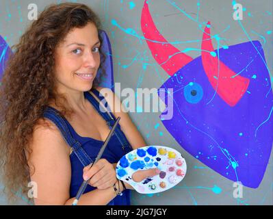 Smiling beautiful young female Mexican painter (visual artist) with long curly hair holds a brush and a palette in front of her painting. Stock Photo