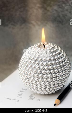 Close-up of beautiful round silver candle burning on sheet of paper with inscription I miss you, hand lettering. Against the background of frosted window glass at night. Letter memories. Black pencil Stock Photo