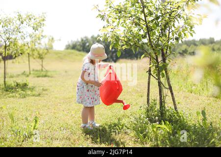 A little cute baby girl 3-4 years old in a dress watering the plants from a watering can in the garden. Kids having fun gardening on a bright sunny Stock Photo