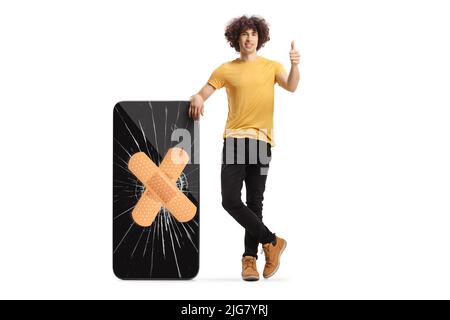 Guy leaning on a broken smartphone fixed with adhesive bandage and showing thumbs up isolated on white background Stock Photo