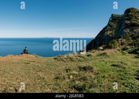 The man sitting on the edge of the green cliff against the blue sea and sky. Madeira, Portugal. Stock Photo