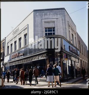 Burton, 16-18 High Street, Abergavenny, Monmouthshire, Wales, 1980s-1990s. The Burton store at 16-18 High Street, showing the carved logo, polished dark stone facade, and chain of merit transom lights. The Burton store in Abergavenny was built in 1937, and a foundation stone was laid by Raymond Montague Burton in that year. The store was designed by their in-house architect Nathaniel Martin. It features common Burton motifs including the logo carved in relief in stone, the 'chain of merit' transom lights at the top of the shopfront windows, and the fascia of polished dark stone. Stock Photo