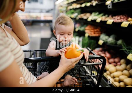 A cute curious baby girl sitting on a trolley and touching a pepper at a store Stock Photo
