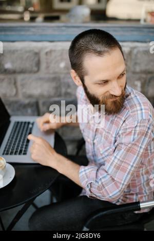 A vertical of a young Caucasian man working remotely from an outdoor cafe Stock Photo