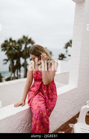 A vertical of a Caucasian female in a cute pink dress posing on a balcony against a beach in Mexico Stock Photo