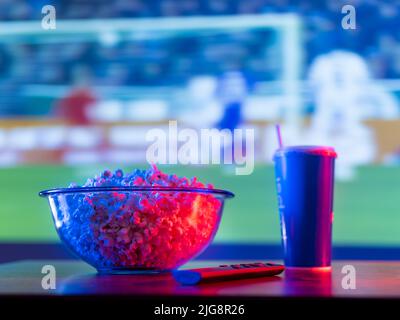 Football match on the big screen plasma TV. On the table is a bowl of popcorn, a soda drink in a plastic cup, and a remote control. Watching sports pr Stock Photo