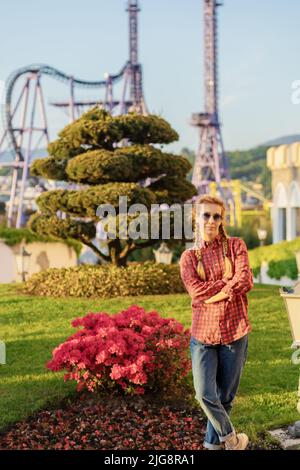 A woman in youth shirt and jeans poses standing near a flower bed with pink flowers and a green decorative tree Stock Photo