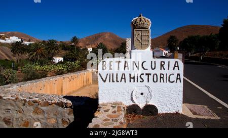 Spain, Canary Islands, Fuerteventura, Betancuria, Old Capital, Old Town, Stone White Town Entrance Sign, Villa Historica, Asphalt Road, Blue Cloudless Sky Stock Photo