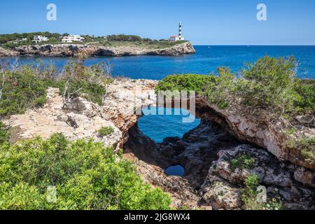 Spain, Balearic islands, Mallorca, district of Felanitx, Portocolom. Sa Cova Foradada, natural arch of rock and in background the lighthouse of Punta de Ses Crestes Stock Photo