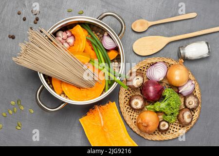 Mushrooms, onions and broccoli on wicker plate. Noodles, pumpkin and chickpeas in saucepan. Flat lay. Grey background. Stock Photo