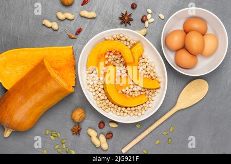 Sliced pieces of pumpkin and chickpeas in white plate. Half a pumpkin and wooden spoon on table. Eggs in bowl. Flat lay. Grey background. Stock Photo