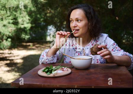 Charming woman sits at rustic dining table, tastes Ukrainian national dish- traditional Borscht with green onion, sliced bacon, parsley and whole grai Stock Photo