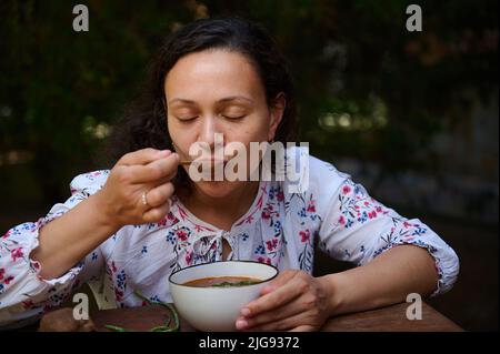 Close-up portrait of a happy multiethnic beautiful woman tasting delicious Ukrainian national dish - traditional Borscht on the nature background. Sla Stock Photo