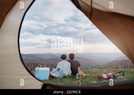 View from inside tent on beautiful landscape of mountains and couple resting. Love and travel.  Stock Photo