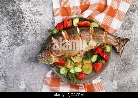Oven baked rainbow trout with vegetables and greens on a round plate on a dark background. Top view, flat lay Stock Photo