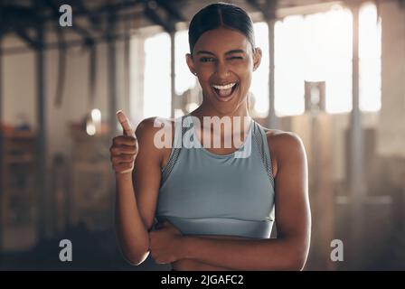 I hope you have a smashing session today. Portrait of a sporty young woman winking while showing thumbs up in a gym. Stock Photo