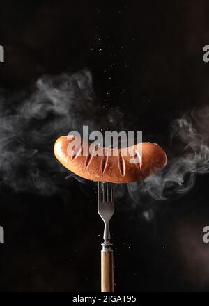 Single tasty roasted grilled sausage withspices sprinkled on rustic fork against dark black background with vapor. Copy space, vertical orientation Stock Photo
