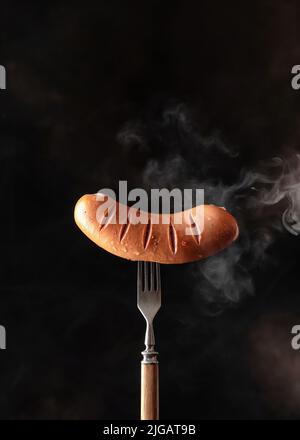 Single tasty roasted grilled sausage with fried stripes on rustic fork against dark black background with vapor. Copy space, vertical orientation Stock Photo