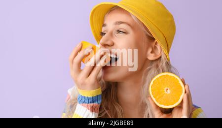 Beautiful young blonde woman eating orange on lilac background Stock Photo