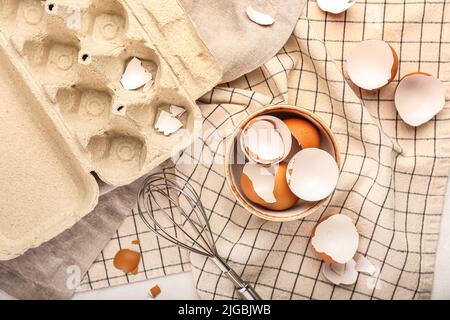 Composition with bowl of broken egg shells, whisk and holder on fabric background Stock Photo