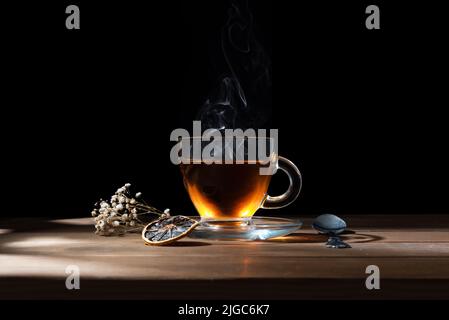 Close-up of a beautiful glass mug with hot tea on a wooden table with dried flowers and dried lemon on black background Stock Photo