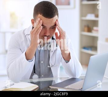 How am I going to tell my patient. a handsome young doctor sitting alone in his clinic and feeling stressed while using his laptop. Stock Photo