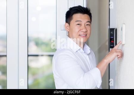 Asian businessman portrait, man walks into modern office, uses doorbell with fingerprint lock, man looks at camera and smiles Stock Photo
