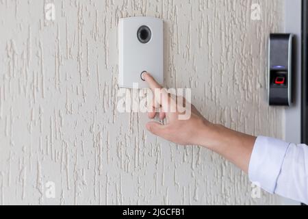 Hand of a man in a white shirt pressing the button of a door knocker on a white wall of a house, a smart door knocker with a video camera for communic Stock Photo