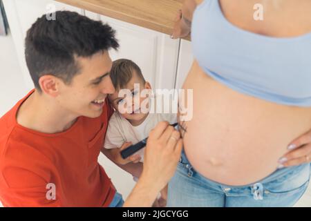 Happy parenting concept. Grinning young adult man and his little son write something on their beloved wife mother belly. High quality photo Stock Photo