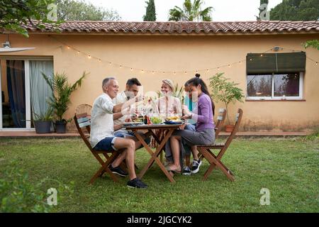 Group of friends gathered around a table in a garden on a summer afternoon to share a meal and have a good time together. Stock Photo