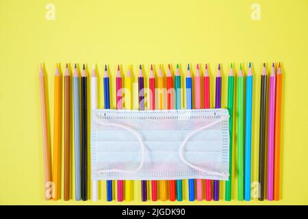 Multicolored colored pencils close-up from above protective mask on yellow background, school time quarantine concept Stock Photo