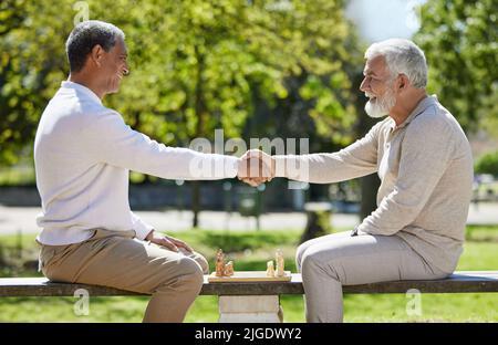 May the best man win. two senior men sitting together on a bench in the park and shaking hands before playing chess. Stock Photo