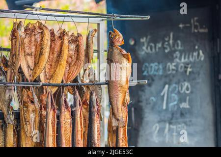 Hanging smoke-dried various fish in a fish market just smoked with hardwood wood chips in a smoker and ready to eat with smoke and info on the board Stock Photo