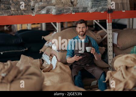 Barista sitting on the warehouse with bags of coffee beans, drinking coffee and using phone Stock Photo