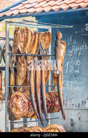 Hanging smoke-dried various fish in a fish market just smoked with hardwood wood chips in a smoker and ready to eat with smoke and info on the board Stock Photo