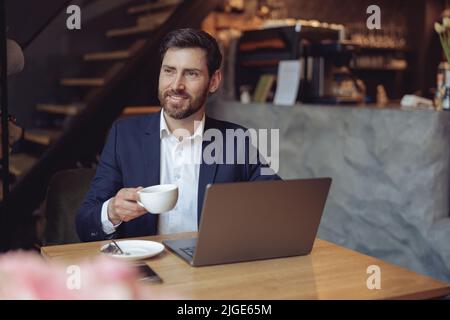 Attractive young elegant businessman holding cup of coffee and smiling at laptop in cafeteria. Stock Photo