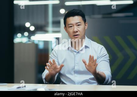 Angry asian businessman talking on video call online meeting, looking at web camera, gesticulating angrily with hands, man at work in office Stock Photo