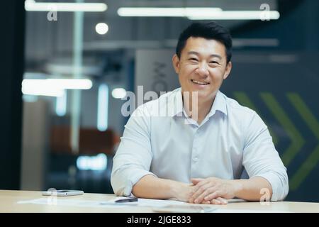 Asian businessman in modern office looking at webcam, smiling sitting at table, online meeting Stock Photo
