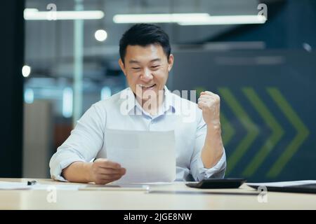 Successful and confident asian businessman reading bank letter and financial report, happy and celebrating triumph success, holding hand up Stock Photo