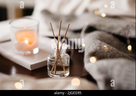 Home liquid fragrance on open paper book and burning scented candle on wooden tray and knit cloth sweater in bed close up. Cozy atmosphere. Stock Photo