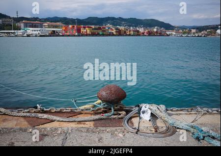 Rusty mooring bollard with ropes and chain on pier in port of La Spezia, Italy. Cargo dock with containers in background Stock Photo