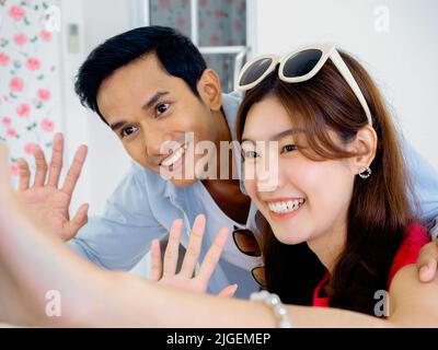 Happy young Asian couple in colorful casual shirts are waving hands in good mood, video call on mobile smartphone screen. Close-up smiling young coupl Stock Photo