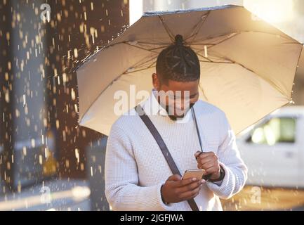 When good news comes pouring in. a young businessman using a cellphone while holding an umbrella on a rainy day in the city. Stock Photo