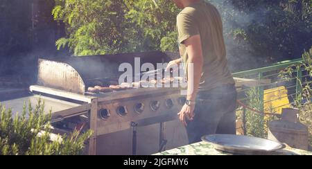 Young guy cooking sausages over open fire while camping in nature. BBQ weekend, grilled pork . Stock Photo