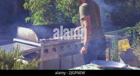 Guy cooking sausages on barbecue grill, open fire on a summer day in the backyard of a private house. Stock Photo