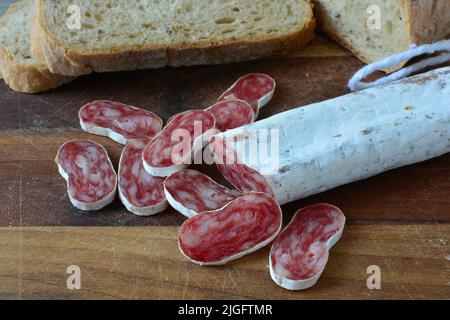 Spanish sausage with white mold, sliced on dark wooden chopping board, with some dark bread in background, close up view Stock Photo