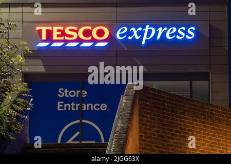 Tesco Express - a small grocery supermarket - logo and sign illuminated at night in The Malls shopping centre in Basingstoke town centre. England, UK Stock Photo