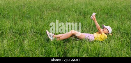 A preteen girl lies on a green grass lawn and takes a selfie on a mobile phone. Horizontal frame Stock Photo