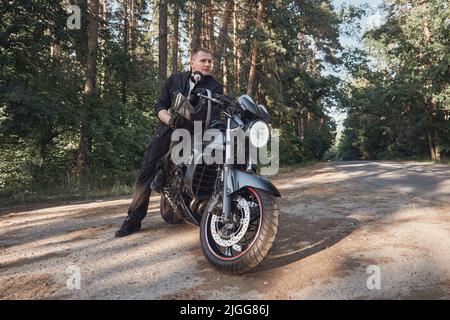Young male biker travels on a motorcycle alone, stopped on the side of a forest road Stock Photo