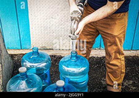 Man in work pants with metal nozzle filling blue bottles with water to use in cabin with no running water - standing beside building on rough backtop Stock Photo